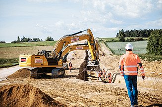 Kotrógép ássa a kábelárkot, az építésvezető figyeli a munkát, a háttérben zöld mező