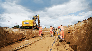 Construction workers compacting a cable trench, yellow excavator in the background, dark clouds in the sky