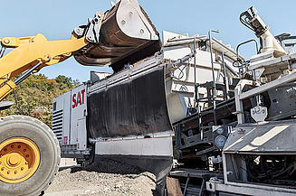 Mobile mixing plant loads recycled material during open-air road construction work.