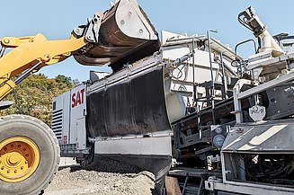 A mobil keverőállomás újrahasznosított anyagot rakod be egy szabadtéri útépítési munkálat során.