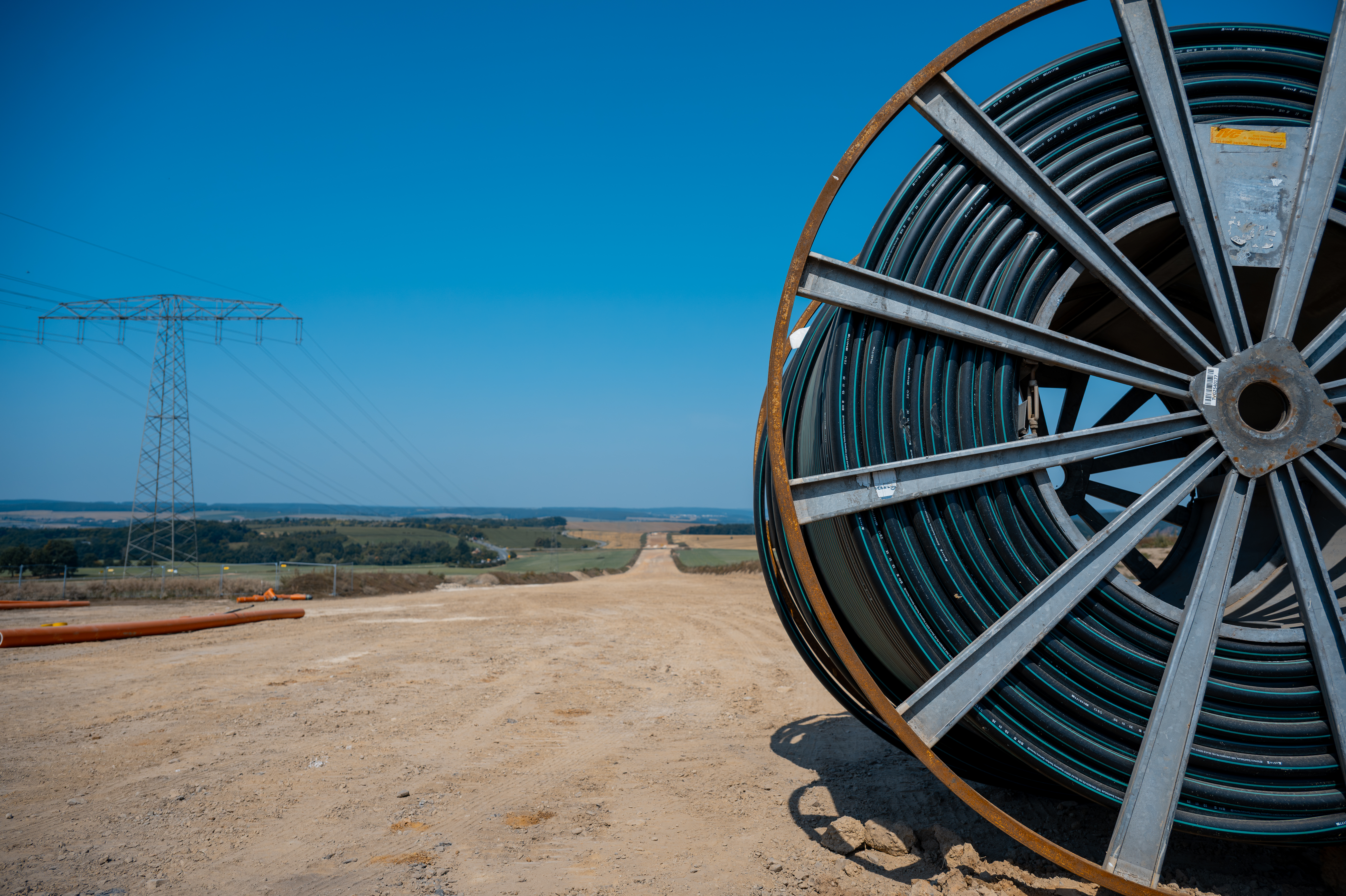 A large cable reel on a construction site for the construction of power lines, with a power pylon and the countryside in the background