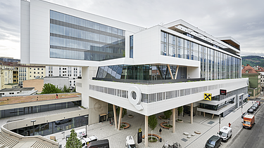 Modern exterior view of the RAIQA quarter in Innsbruck featuring a distinctive glass façade and open ground-floor area. 