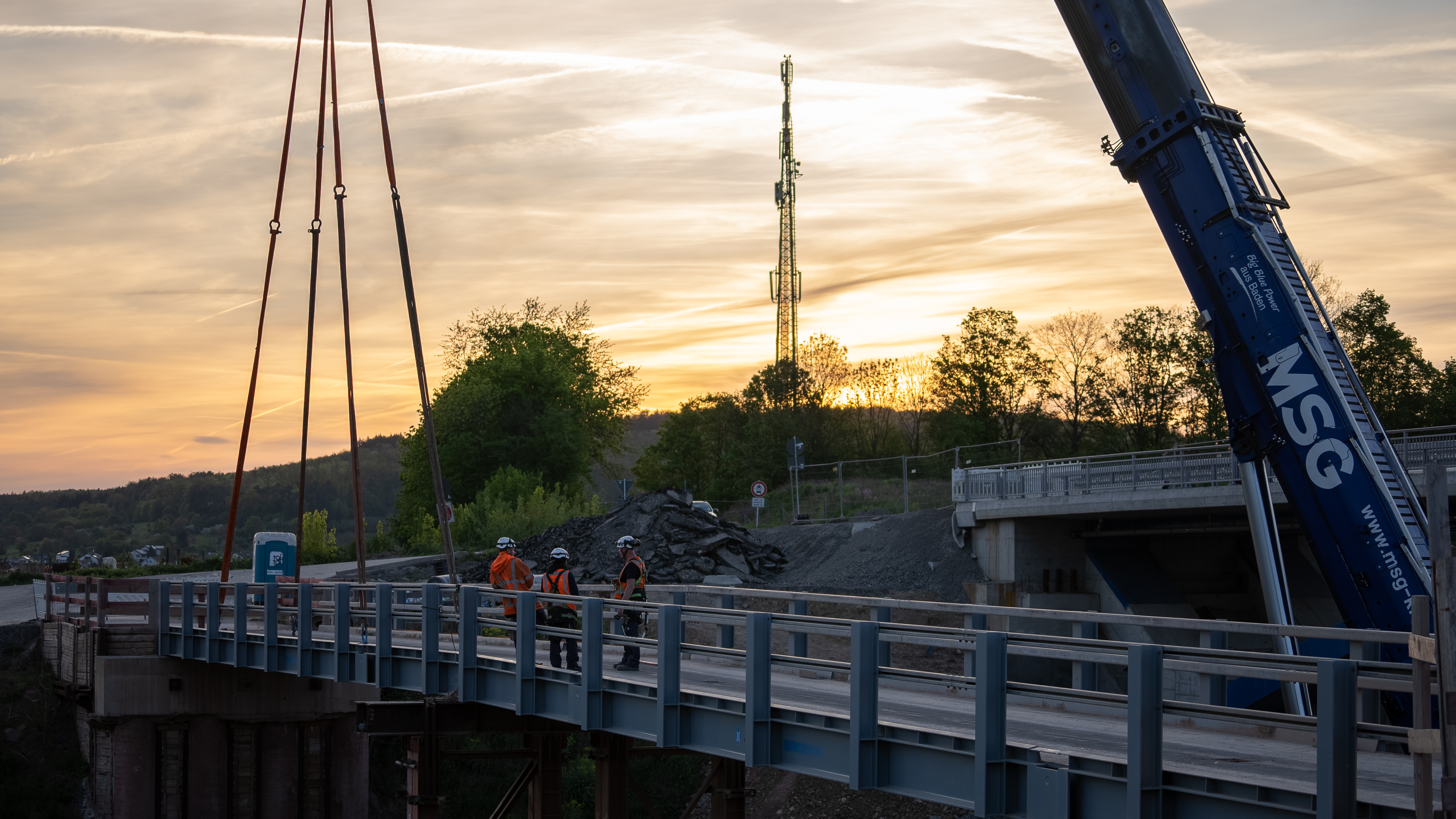 Montage einer Stahlbrücke mit Autokran und Baupersonal bei Sonnenuntergang