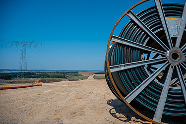 Große Kabeltrommel auf Baustelle für Stromtrassenbau, im Hintergrund Strommast und Landschaft