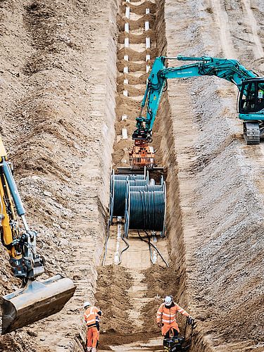 Photo of cables being laid in a deep trench with excavators and construction workers