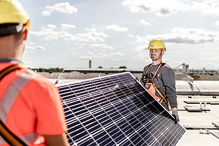 Fotografie a unor muncitori în construcții care transportă un panou solar pe care se văd clar celulele solare. 