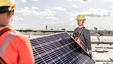 Fotografie a unor muncitori în construcții care transportă un panou solar pe care se văd clar celulele solare. 