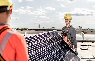 Zdjęcie pracowników budowlanych niosących panel słoneczny, na którym wyraźnie widać ogniwa słoneczne. 