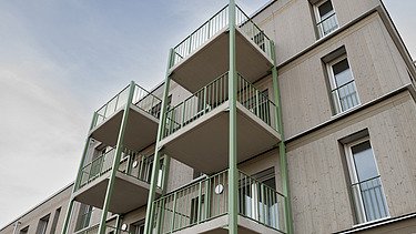 Photograph of a multi-storey residential building constructed using a standardised timber-frame method, featuring green balcony structures